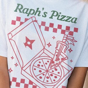 May include: White t-shirt with a red and white illustration of a pizza box and a slice of pizza. The text "Raph's Pizza" is printed in green on the shirt. The text "Wise man say forgiveness is divine, never pay full price for late pizza" is printed in green at the bottom of the shirt.