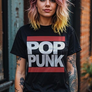 May include: Black t-shirt with the words "POP PUNK" in white block letters, framed by red lines. The person is wearing blue jeans and has colorful hair. The background includes a brick wall and a weathered metal surface.