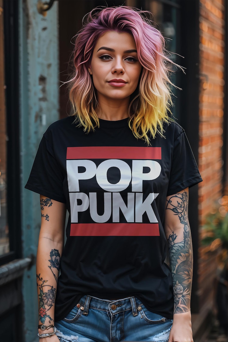May include: Black t-shirt with the words "POP PUNK" in white block letters, framed by red lines. The person is wearing blue jeans and has colorful hair. The background includes a brick wall and a weathered metal surface.