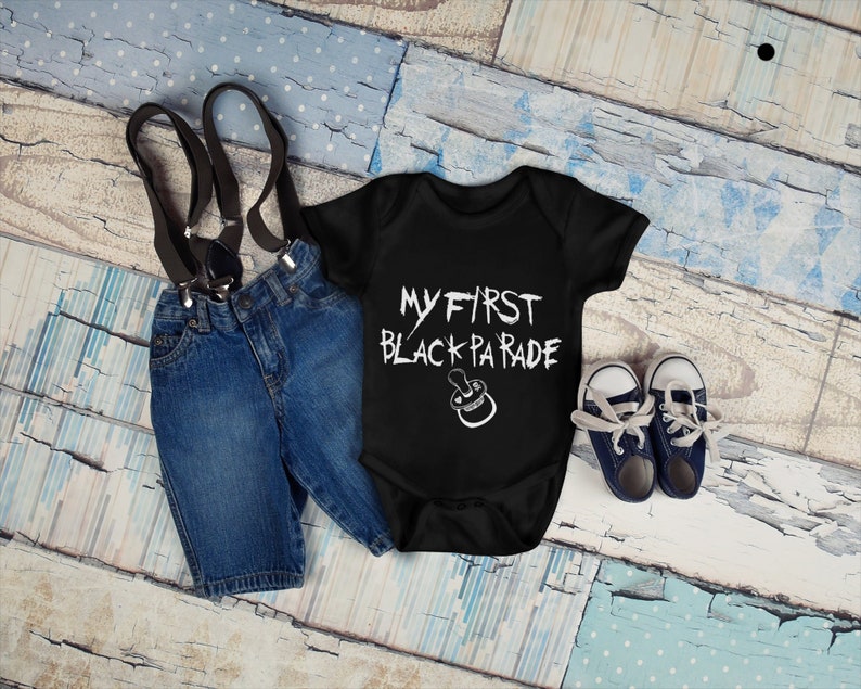 May include: Black bodysuit with white text that reads "My First Black Parade" and a pacifier graphic. The bodysuit is paired with blue denim shorts and black suspenders.