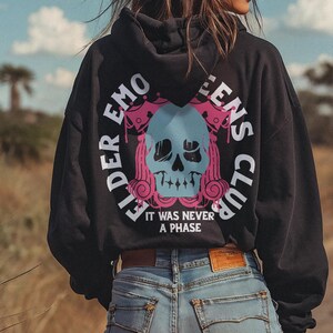Pu&ograve; includere: Felpa con cappuccio nera con un grafico di un teschio e il testo "Elder Emo Queens Club" e "It was never a phase". La modella indossa la felpa con pantaloncini di jeans blu.