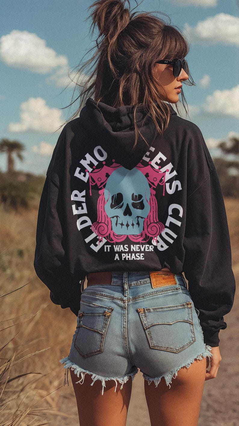 Pu&ograve; includere: Felpa con cappuccio nera con un grafico di un teschio e il testo "Elder Emo Queens Club" e "It was never a phase". La modella indossa la felpa con pantaloncini di jeans blu.