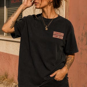 May include: Black t-shirt with the text "COWGIRLS FROM HELL" in red and white. The shirt is worn with black shorts. The image is taken outdoors in front of a building with a "VACANCY" sign.