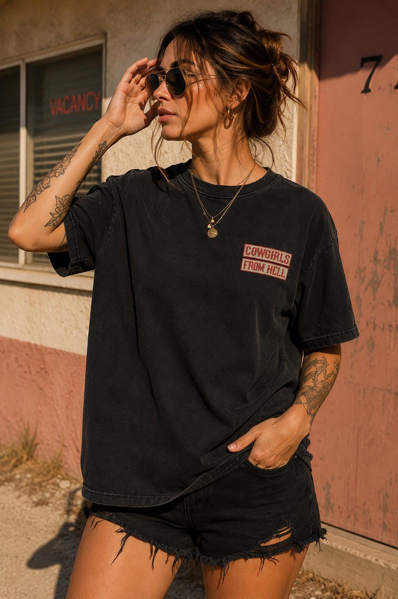 May include: Black t-shirt with the text "COWGIRLS FROM HELL" in red and white. The shirt is worn with black shorts. The image is taken outdoors in front of a building with a "VACANCY" sign.