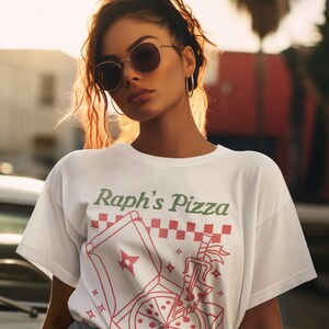 May include: White t-shirt with a graphic of a pizza box and a pizza slice with the text "Raph's Pizza" in green and red lettering.
