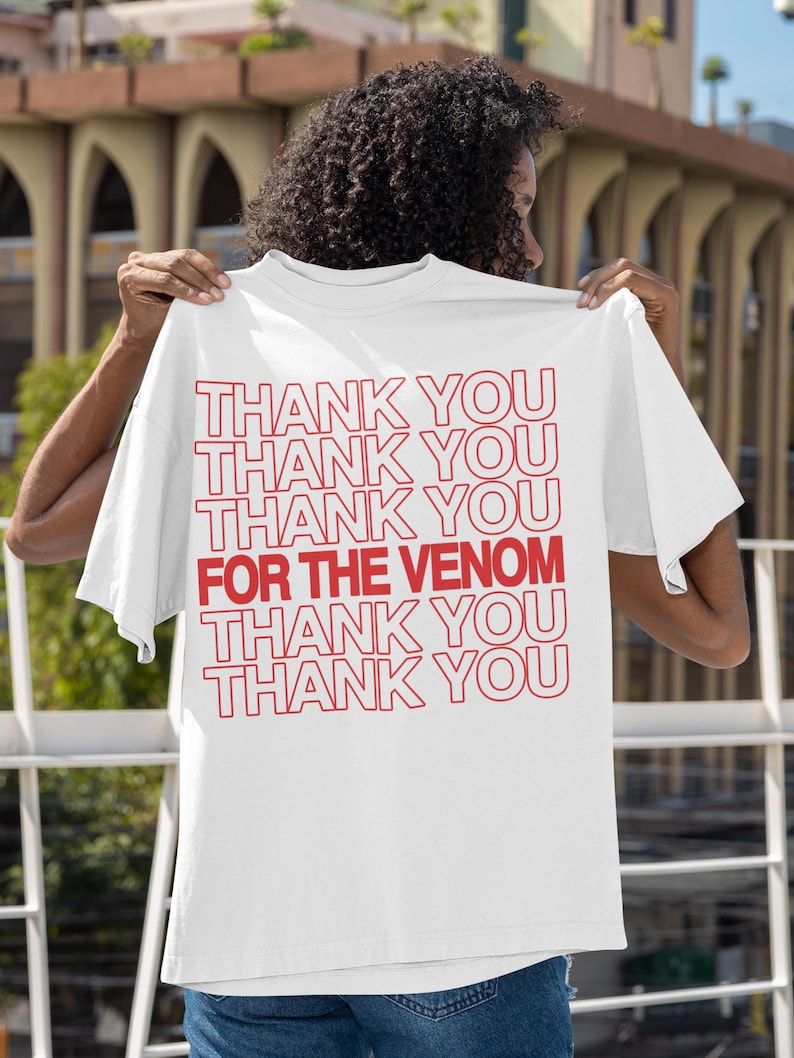 Pu&ograve; includere: Maglietta bianca con testo rosso che recita "THANK YOU" ripetuto pi&ugrave; volte, con "FOR THE VENOM" in un carattere rosso pi&ugrave; audace. La maglietta &egrave; mostrata su uno sfondo sfocato all'aperto.