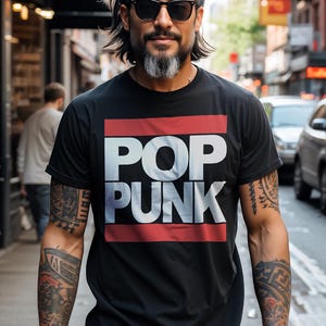 May include: Black t-shirt with the words "POP PUNK" in white block letters, framed by red stripes. The person wearing the shirt has tattoos on both arms and is wearing sunglasses. The background is a city street.