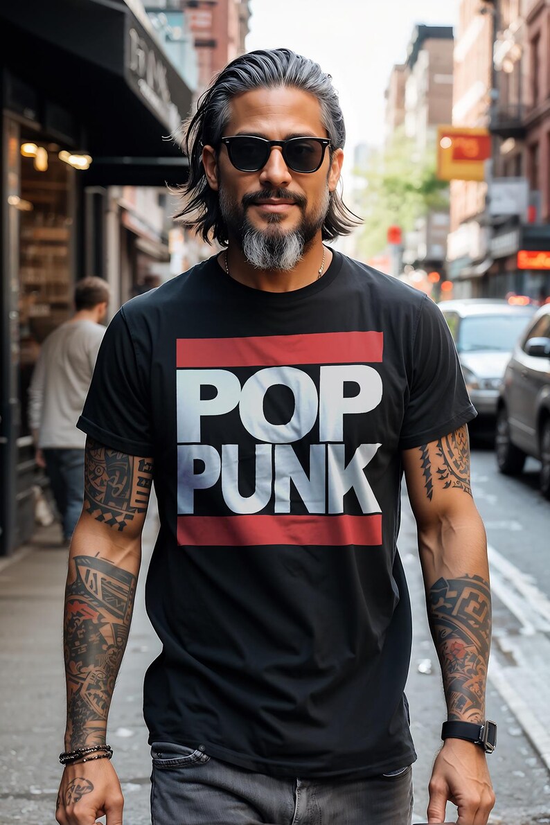 May include: Black t-shirt with the words "POP PUNK" in white block letters, framed by red stripes. The person wearing the shirt has tattoos on both arms and is wearing sunglasses. The background is a city street.