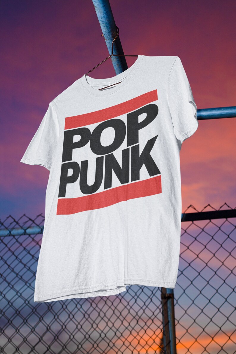 May include: White t-shirt with the words "POP PUNK" in black block letters, framed by red horizontal stripes. The shirt hangs on a hanger against a backdrop of a chain-link fence and a colorful sunset.