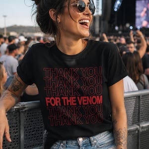Pu&ograve; includere: T-shirt nero con la scritta "THANK YOU THANK YOU THANK YOU FOR THE VENOM THANK YOU THANK YOU" in rosso. La maglietta &egrave; indossata con pantaloncini di jeans blu. La persona sorride e indossa occhiali da sole.