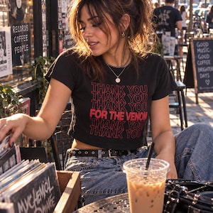 May include: A black short-sleeved crop top featuring the repeated red text THANK YOU and the phrase FOR THE VENOM printed in a bold font on the front. The garment is paired with blue denim jeans and a black belt with silver-toned metal eyelets.