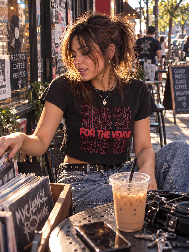 May include: A black short-sleeved crop top featuring the repeated red text THANK YOU and the phrase FOR THE VENOM printed in a bold font on the front. The garment is paired with blue denim jeans and a black belt with silver-toned metal eyelets.