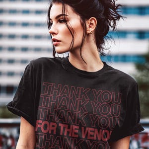 Pu&ograve; includere: T-shirt nera con la scritta "THANK YOU" ripetuta pi&ugrave; volte in rosso. Anche la frase "FOR THE VENOM" &egrave; in rosso. La maglietta &egrave; indossata con jeans blu.