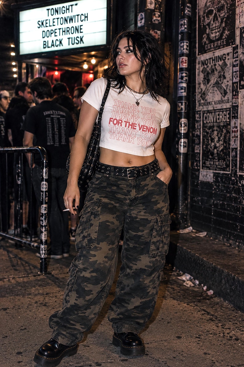 May include: A white short-sleeved crop top featuring the repeated text THANK YOU in light pink and the phrase FOR THE VENOM in bold red lettering, paired with loose-fitting camouflage cargo pants and a black studded leather belt.