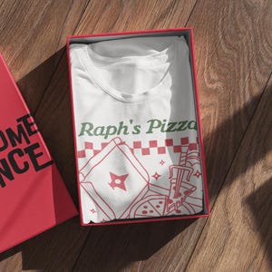 May include: White t-shirt with a red and green graphic of a pizza box with a ninja star and the text "Raph's Pizza".