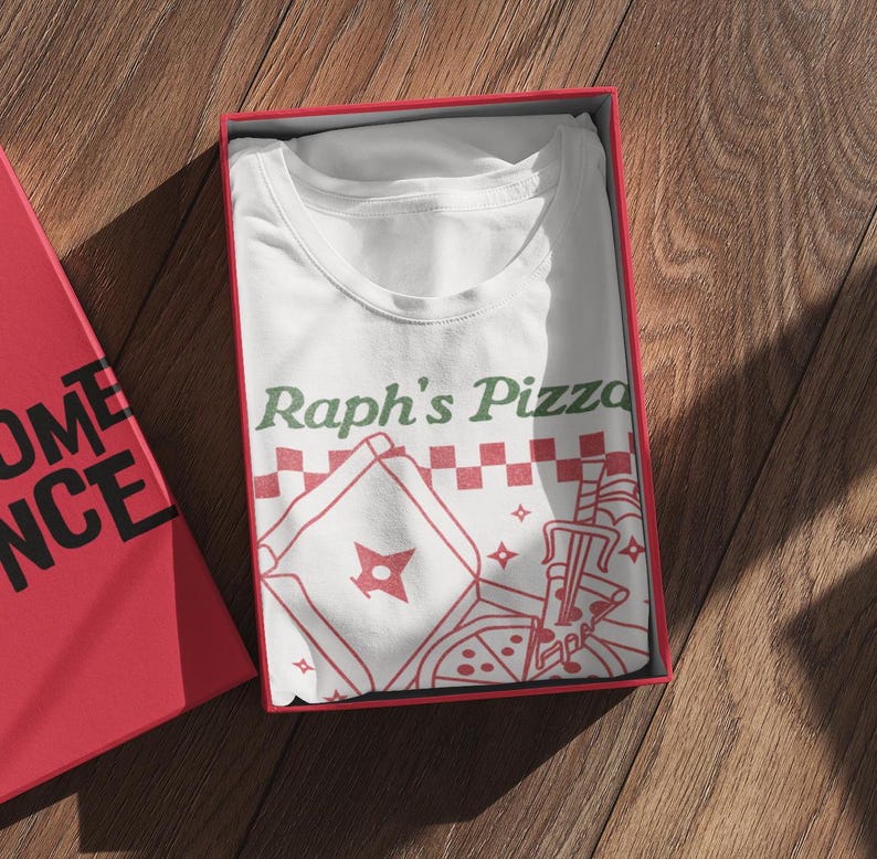 May include: White t-shirt with a red and green graphic of a pizza box with a ninja star and the text "Raph's Pizza".