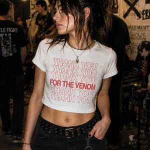 May include: A white short-sleeved crop top featuring the repeated red text THANK YOU with the phrase FOR THE VENOM highlighted in bold red lettering across the center. The garment is paired with black pants and a black belt with metal grommets.