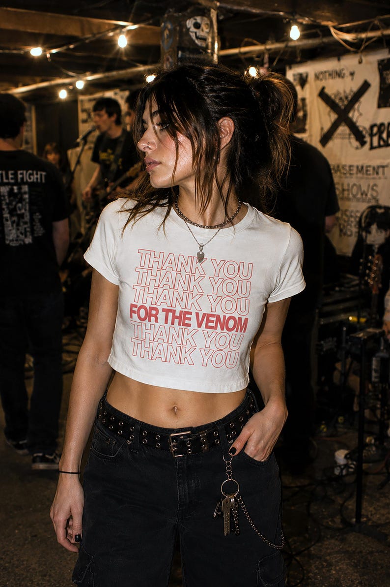 May include: A white short-sleeved crop top featuring the repeated red text THANK YOU with the phrase FOR THE VENOM highlighted in bold red lettering across the center. The garment is paired with black pants and a black belt with metal grommets.