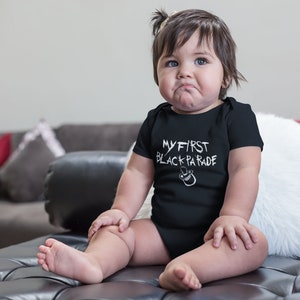 May include: A baby wearing a black bodysuit with the text "MY FIRST BLACK PARADE" in white. The baby is sitting on a black leather couch with a white fur throw pillow.