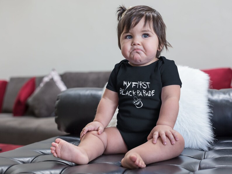 May include: A baby wearing a black bodysuit with the text "MY FIRST BLACK PARADE" in white. The baby is sitting on a black leather couch with a white fur throw pillow.