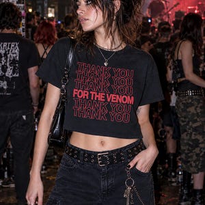 May include: A black cropped t-shirt featuring the repeated red text THANK YOU and the phrase FOR THE VENOM in a larger font. The shirt is paired with black cargo pants, a black leather belt with silver grommets, and a silver chain accessory.