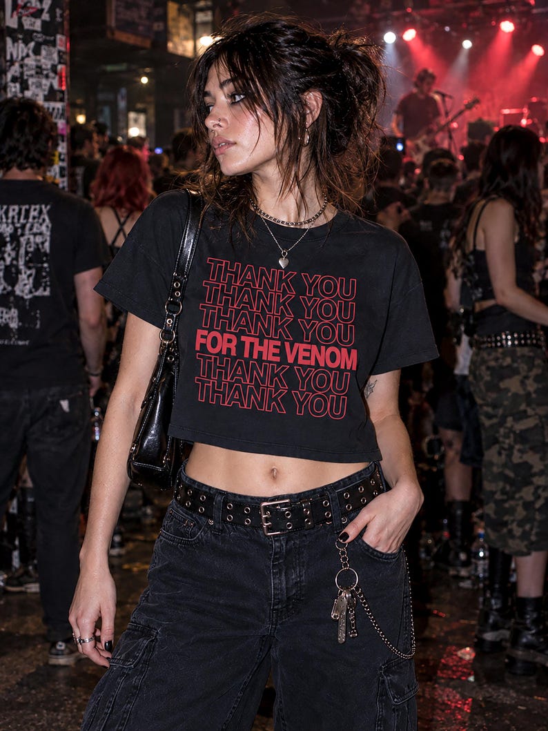 May include: A black cropped t-shirt featuring the repeated red text THANK YOU and the phrase FOR THE VENOM in a larger font. The shirt is paired with black cargo pants, a black leather belt with silver grommets, and a silver chain accessory.