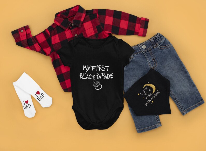 May include: A black baby onesie with the text "My First Black Parade" in white lettering. The onesie is laid out on a yellow surface with a red and black plaid shirt, blue jeans, and two white socks with red hearts and the text "I love Dad" on them.