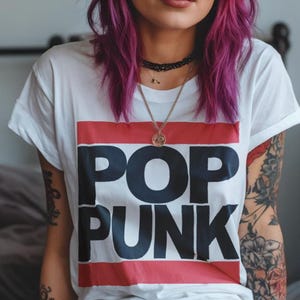 May include: A person wearing a white t-shirt with the words "POP PUNK" in large black letters, set against a red and blue stripe. The person has vibrant purple hair, sunglasses, and tattoos on their arms.