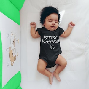 May include: A baby wearing a black onesie with the text "MY FIRST BLACK PARADE" in white. The baby is lying on a white sheet in a crib.
