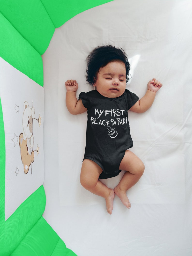 May include: A baby wearing a black onesie with the text "MY FIRST BLACK PARADE" in white. The baby is lying on a white sheet in a crib.