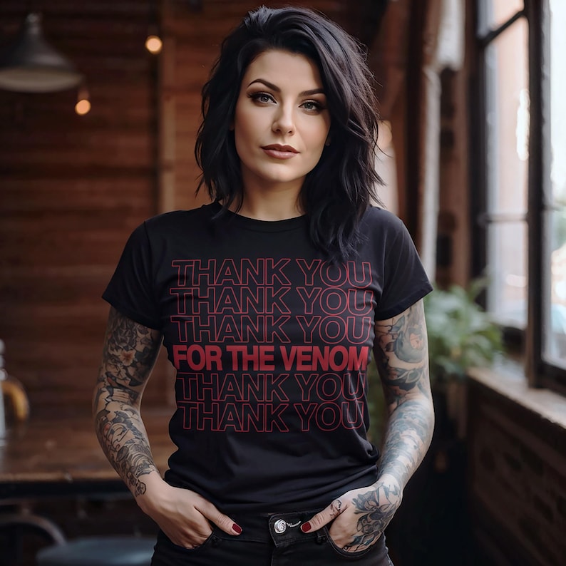 Pu&ograve; includere: T-shirt nero con la scritta rossa "THANK YOU FOR THE VENOM". La maglietta &egrave; a girocollo ed &egrave; indossata da una persona con capelli scuri e tatuaggi visibili sulle braccia.