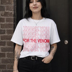 Pu&ograve; includere: T-shirt bianco con la scritta rossa "THANK YOU THANK YOU THANK YOU FOR THE VENOM THANK YOU THANK YOU THANK YOU". La maglietta &egrave; abbinata a jeans neri e una cintura nera.