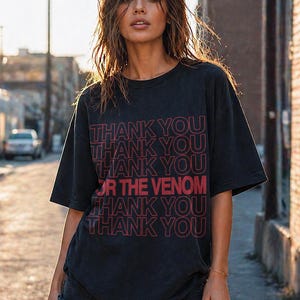 Pu&ograve; includere: T-shirt nero con la scritta "THANK YOU OR THE VENOM" in rosso. La maglietta ha un girocollo e maniche corte. La modella indossa pantaloncini di jeans neri.