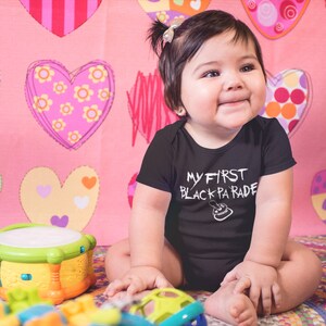May include: A baby wearing a black onesie with the text "My First Black Parade" in white. The baby is sitting on a pink and white heart patterned blanket with a toy drum and stacking rings.