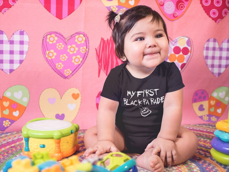 May include: A baby wearing a black onesie with the text "My First Black Parade" in white. The baby is sitting on a pink and white heart patterned blanket with a toy drum and stacking rings.