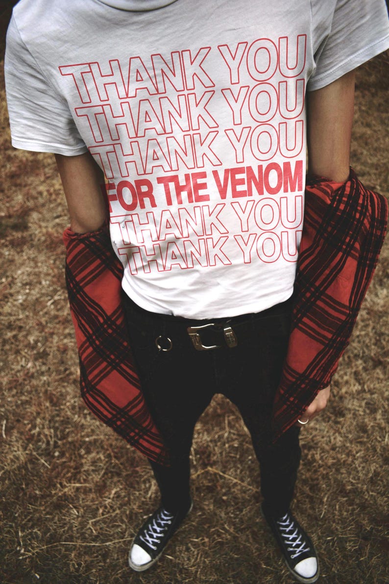 May include: A white t-shirt with the text 'Thank You' repeated multiple times in red. The text 'For The Venom' is also written in red. The shirt is worn with black jeans and a red and black plaid shirt. The person is wearing black and white sneakers.