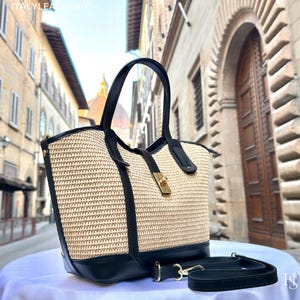 May include: A woven straw tote bag with black leather accents. The bag features a gold-colored clasp and a detachable black shoulder strap. The bag is sitting on a white surface, with a blurred background of a European street.