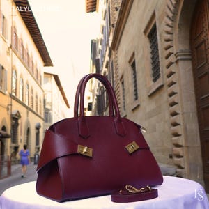 Peut inclure: Un sac à main en cuir bordeaux profond à la surface texturée, doté de deux anses et de fermetures en métal doré sur les côtés. Le sac présente une silhouette structurée et comprend une bandoulière amovible posée sur la surface.