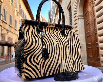 May include: A black and tan zebra print handbag with a black crocodile embossed leather trim and a gold lock. The bag has a black adjustable strap and is photographed on a purple surface.