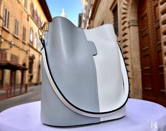 Può includere: Una borsa a secchiello dal design bicolore in pelle martellata grigia e bianca. La borsa presenta una tracolla larga con bordi neri, dettagli in metallo color oro e un'apertura superiore curva e liscia.