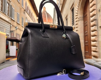 May include: Black leather handbag with gold hardware and a detachable shoulder strap. The bag has a top handle and a lock closure. ITALYLEATHERIT is written on the top of the bag.