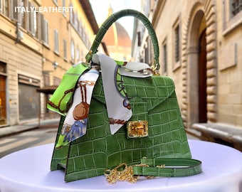 Puede incluir: Un bolso de mano de cuero verde con estampado de cocodrilo, con una correa de cadena dorada y una bufanda estampada en verde y blanco. El bolso tiene un asa superior y una cerradura dorada.