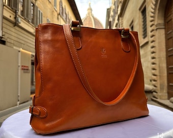 May include: A brown leather tote bag with a gold buckle and a logo that says "Firenze Italy". The bag is sitting on a white surface with a blurry background of a city street.