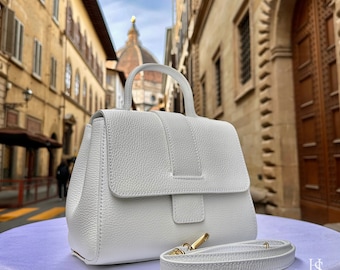 May include: A white leather handbag with a top handle and a detachable shoulder strap. The bag has a flap closure with a gold buckle. The bag is photographed against a backdrop of a city street.