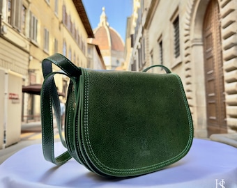 May include: A dark green leather crossbody bag with a flap closure and a long adjustable strap. The bag is photographed in front of a building with a tiled facade.