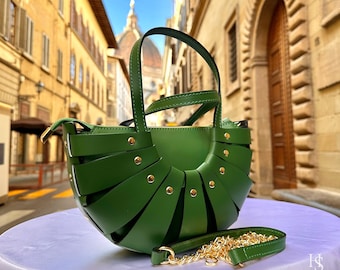 May include: A green leather handbag with a unique woven design. The bag features a top handle and a detachable gold chain strap. Gold-colored rivets accent the woven pattern. The bag is set against a blurred background of a European street.