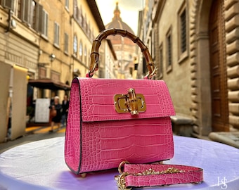 May include: A pink crocodile embossed handbag with a bamboo handle and gold hardware. The bag has a small, rectangular shape and a detachable strap. The bag is sitting on a white surface with a blurry background of a city street.