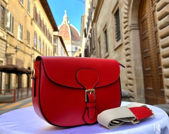 May include: A red leather satchel bag with a gold buckle closure and a beige and red adjustable strap. The bag is sitting on a white surface with a blurry background of an Italian city street.