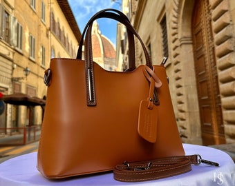 May include: A brown leather handbag with a top handle and a detachable shoulder strap. The bag has a silver metal tag with the text "ITALY LEATHERIT" on it.