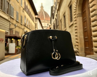 May include: A black leather crossbody bag with a gold-toned clasp and two rings. The bag is sitting on a white surface with a blurry background of a city street.